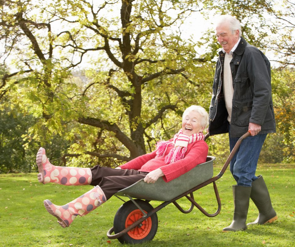 An older man laughs as he pushes his wife across the lawn in a wheelbarrow, her polka-dot wellington boots kicked up in the air