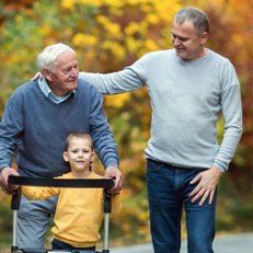 Three generations walk an autumn path together: a grandfather steadying himself on a rollator, a small boy beside him and his adult son's hand on his shoulder