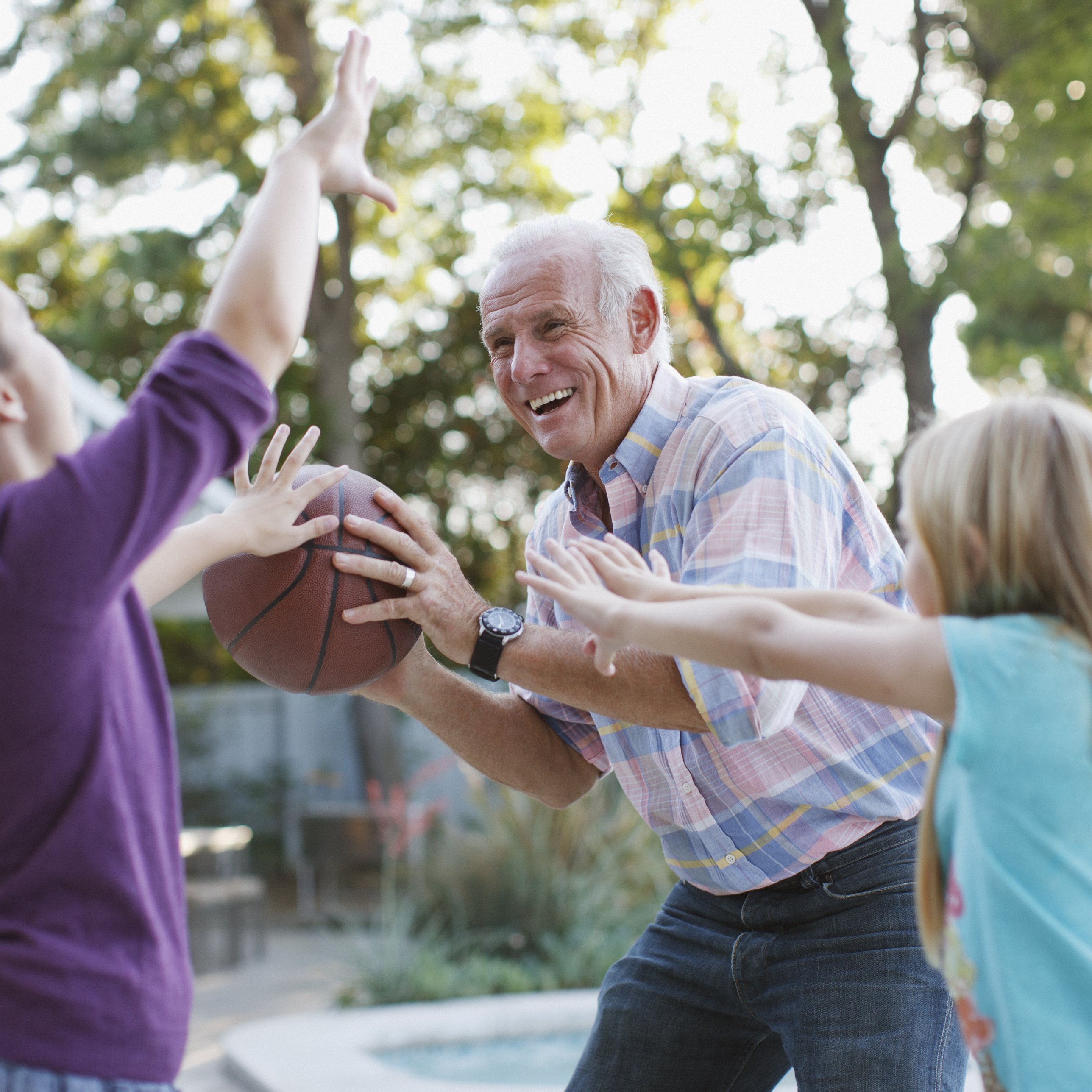 A smiling older man plays basketball with two girls beside a backyard pool