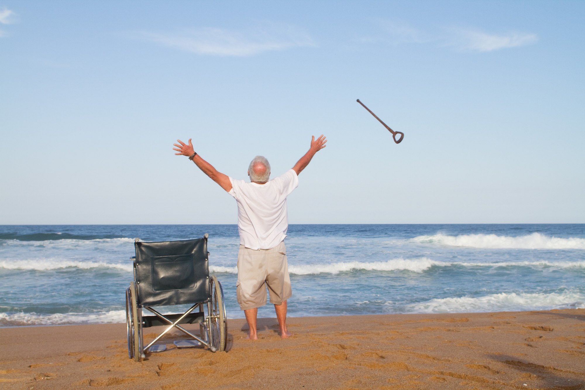 An older man stands barefoot on a beach with both arms raised, his empty wheelchair beside him and his cane flying through the air