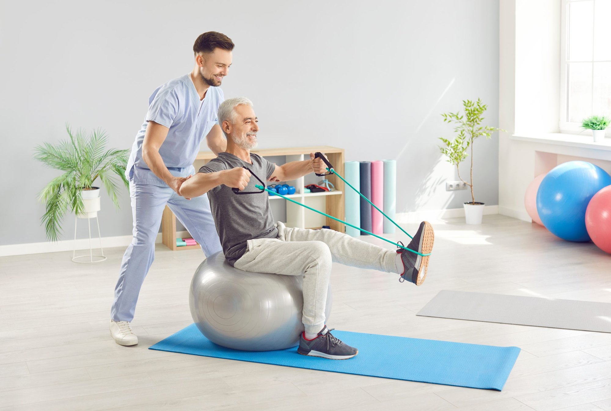 A physical therapist helps an older man exercise on a stability ball with resistance bands